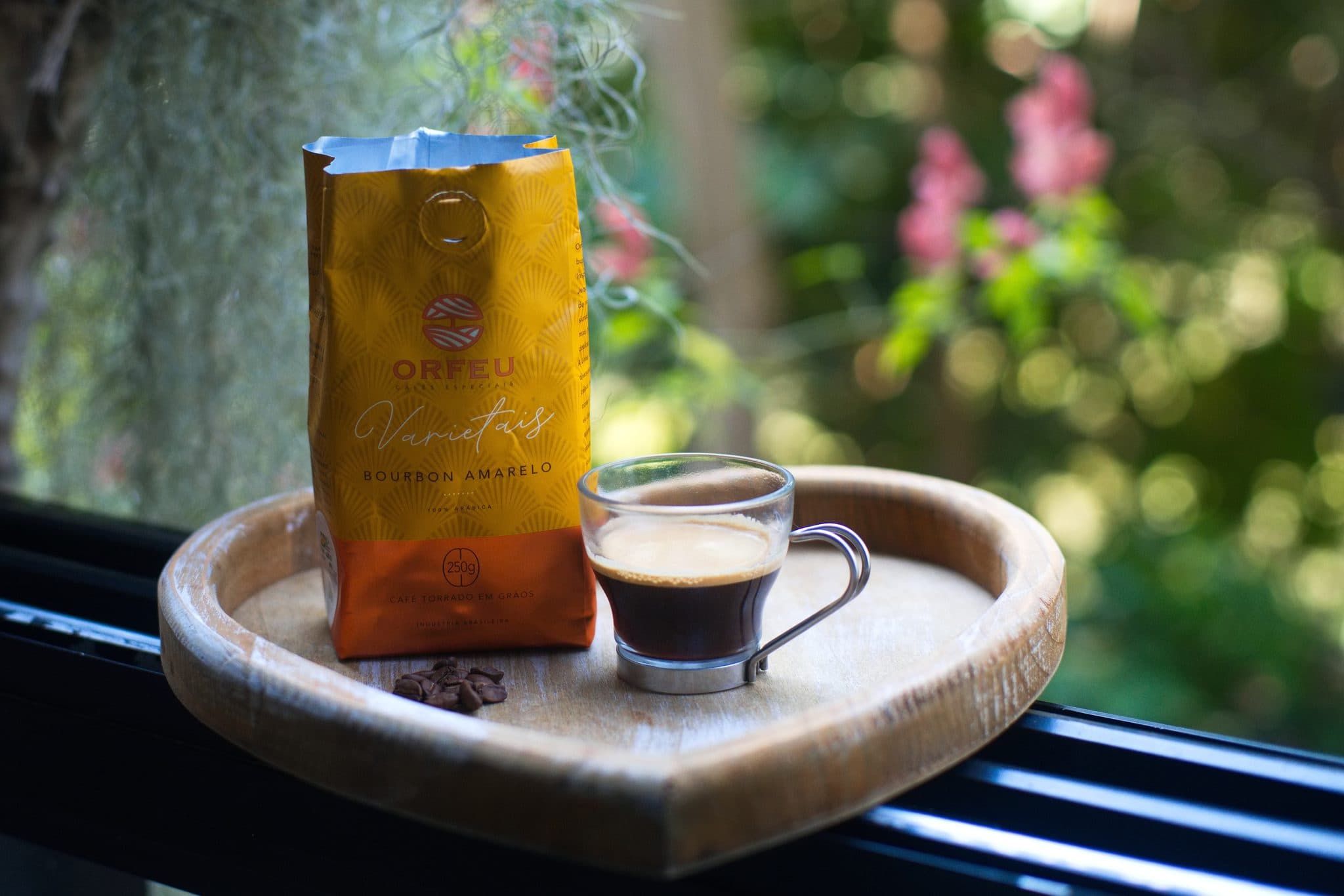 Pacote de 250 g do Orfeu Varietais Bourbon Amarelo e uma xícara de café sobre bandeja de madeira na janela, com o jardim ao fundo