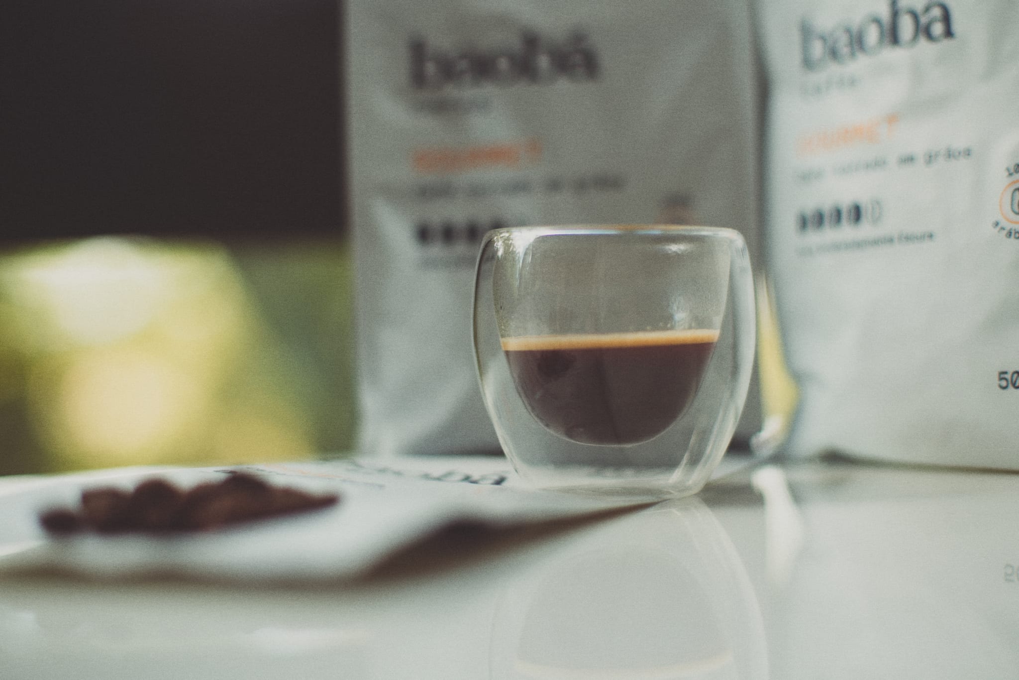 Xícara de vidro de parede dupla com o café Baobá Gourmet em close, mostrando o creme da bebida, com os pacotes do café desfocados ao fundo